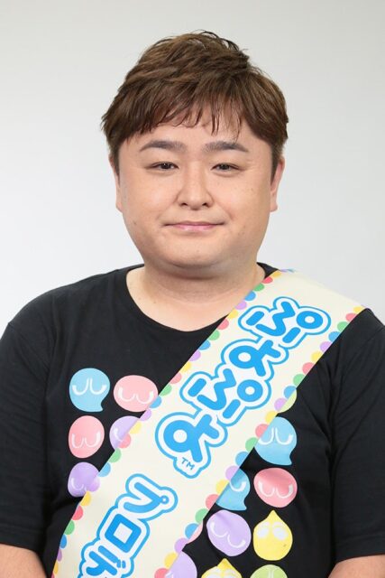 記事画像4: 黒いTシャツを着て、肩に「ぷよぷよ」と書かれたたすきをかけた男性