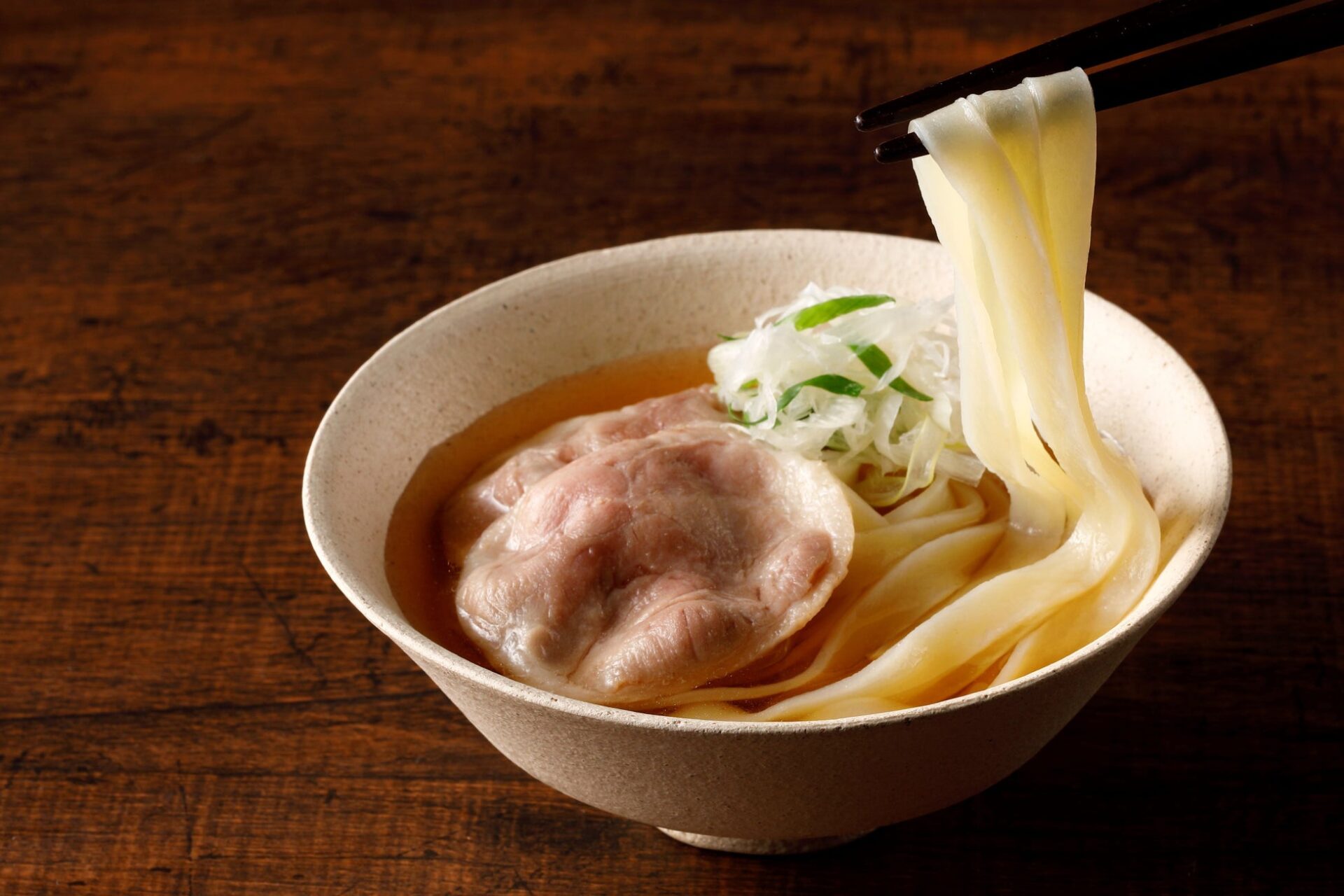 記事画像11: 器に入ったおうどんに、肉とネギが添えられ、箸で麺を持ち上げている様子。
