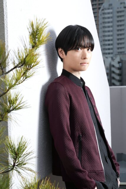 黒髪の男性がワインレッドのジャケットを羽織り、壁にもたれている。