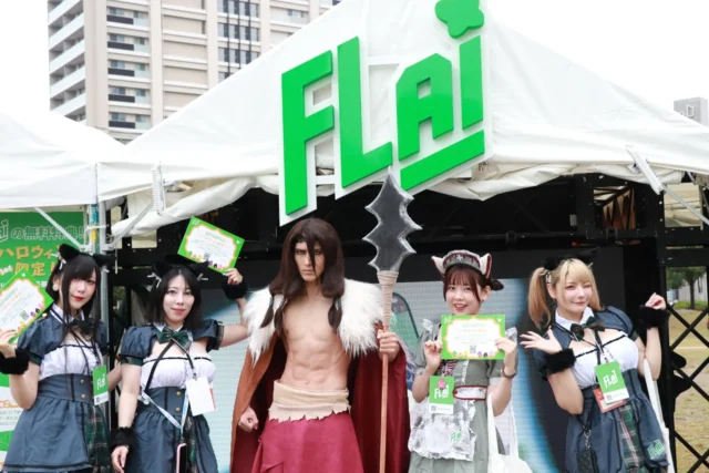 FLAIブース前で笑顔でポーズをとる複数のコスプレイヤーたち