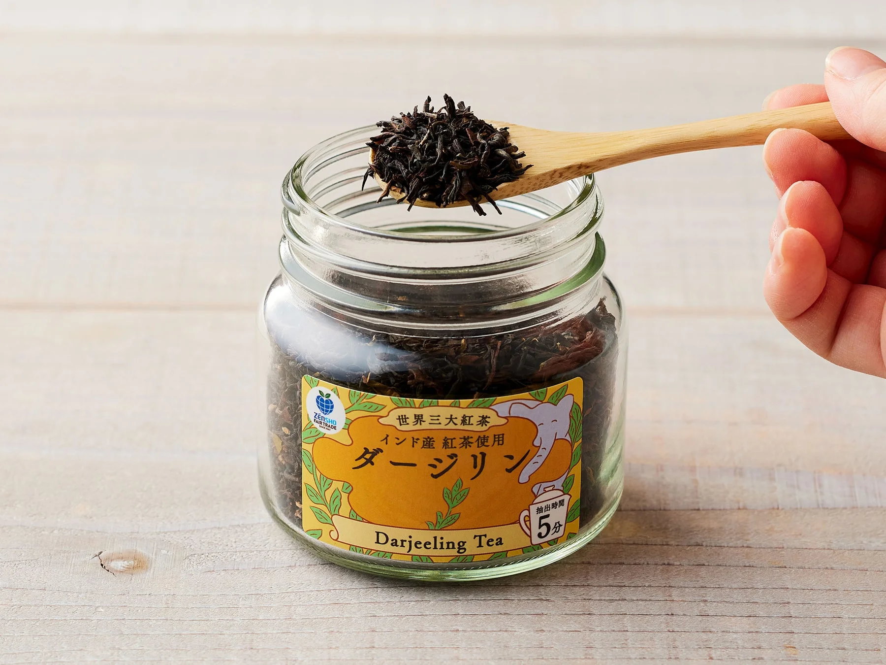 スプーンで掬われたダージリン茶葉が入った瓶。世界三大紅茶の表示とフェアトレードロゴがある