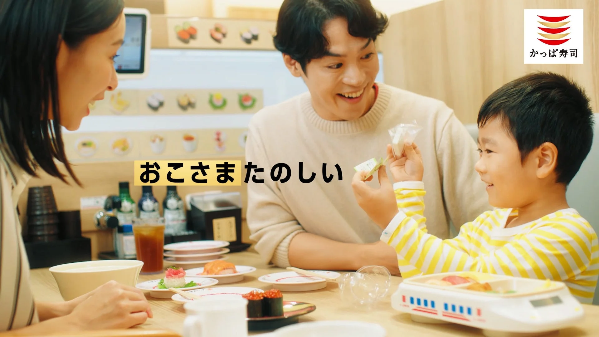 かっぱ寿司のテーブルで子供が笑顔でお菓子を受け取るCMのワンシーン。「おこさまたのしい」の文字が表示されている。