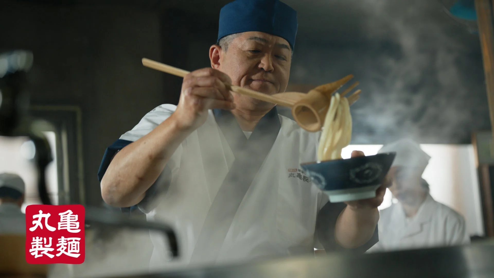 湯気が立ちのぼる釜から打ち立てのうどんをすくい上げる丸亀製麺の麺職人