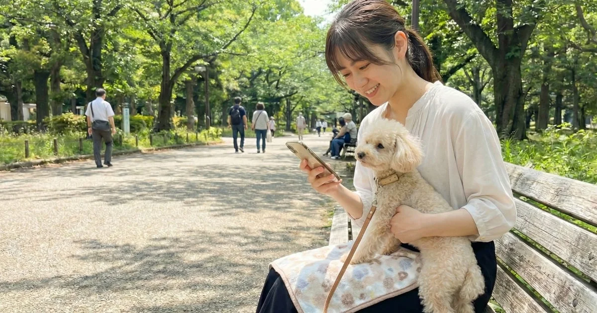 公園のベンチで愛犬を抱きながらスマホを操作する女性。アプリでのおでかけ先探しをイメージさせる。