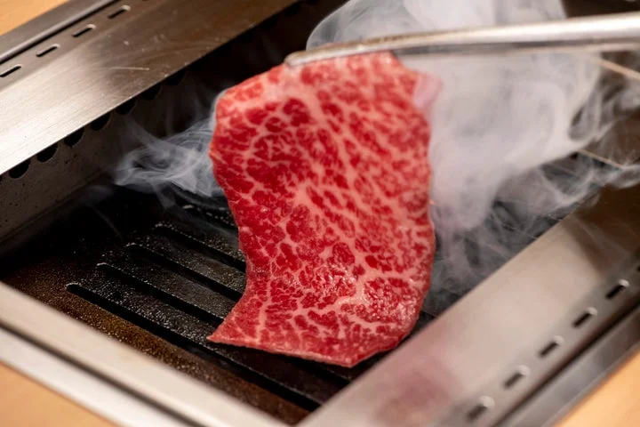 トングで掴まれたA5黒毛和牛が網の上で煙を上げながら焼かれる様子