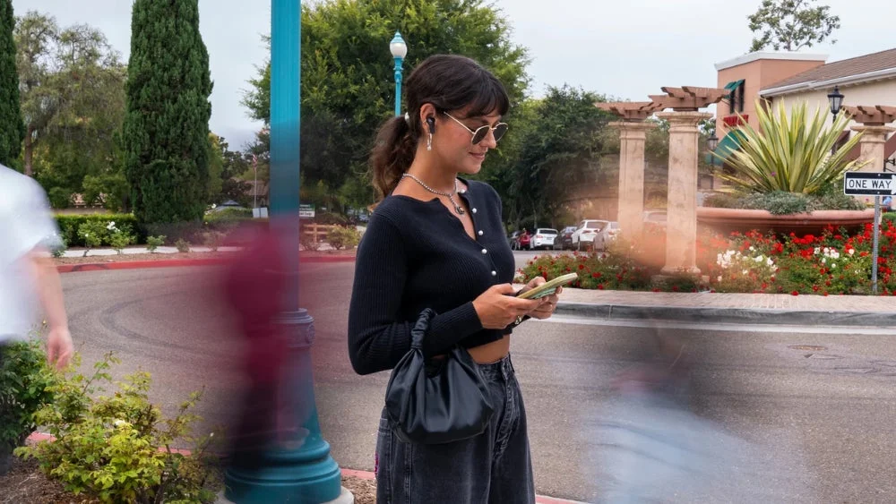 屋外でEpic Pods ANCワイヤレスイヤホンを使用しスマホを操作する女性。