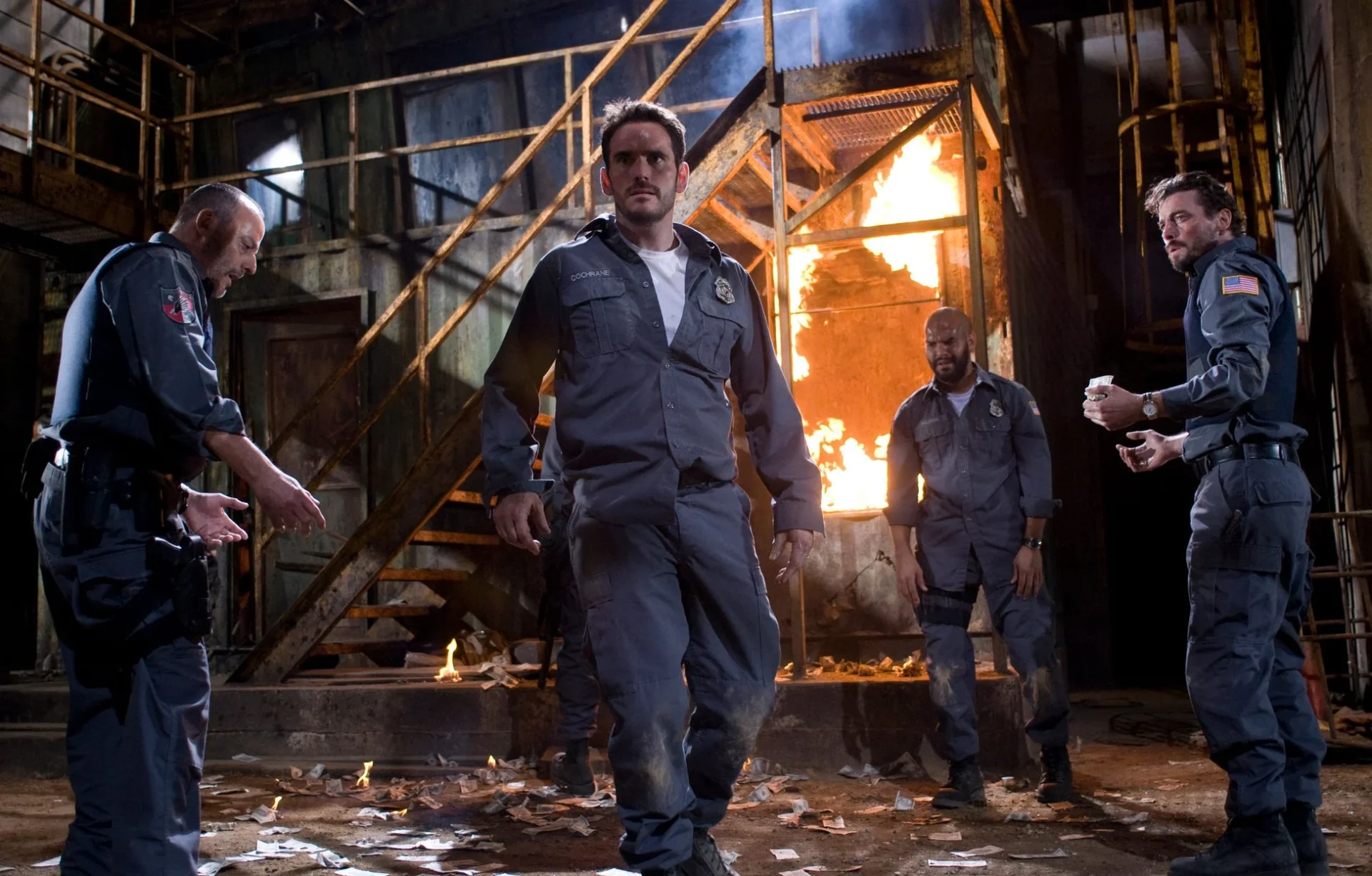 映画『アーマード 武装地帯』のワンシーン。炎と散らばる紙幣の中で困惑する男たち