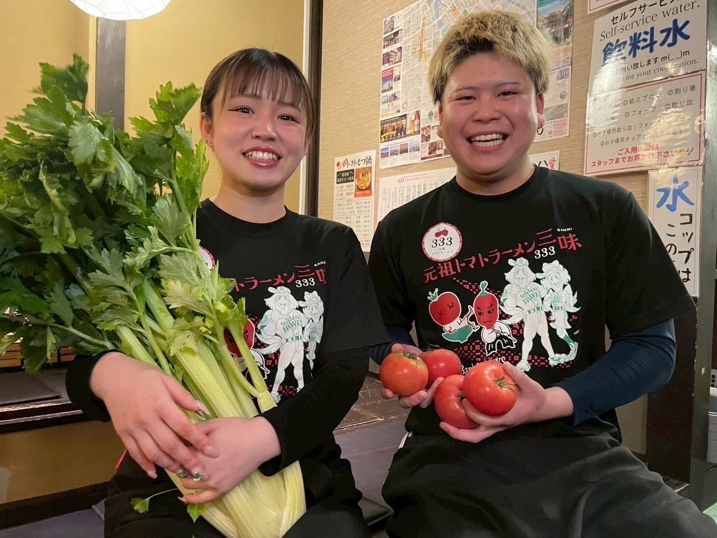 元祖トマトラーメン三味のスタッフがトマトとセロリを持って笑顔で立つ様子
