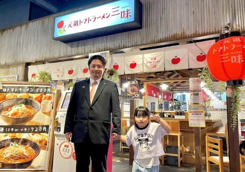 元祖トマトラーメン三味の創業者宮下将司氏と子供が店舗前で手をつないで立つ姿