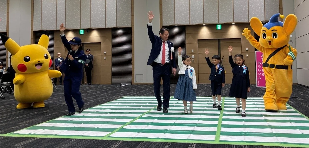 ピカチュウとピーポくん、警察官、児童が交通安全教室で手を挙げる様子