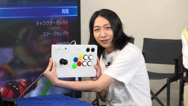 JIMさんがアーケードコントローラーを持って笑顔を見せる